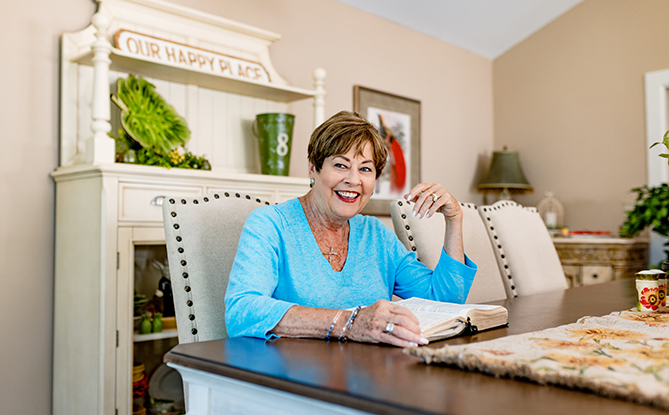 Shelia sitting at her dining room table with an open book, smiling wide